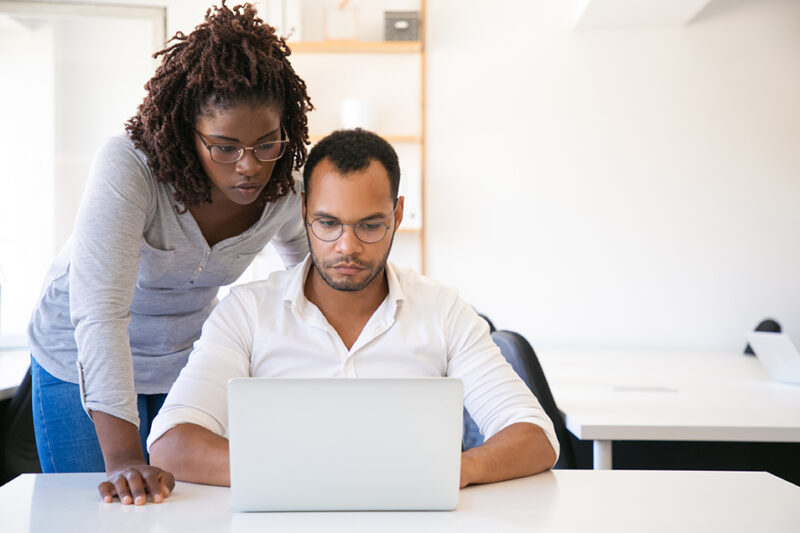 Diverse colleagues watching presentation on computer. Business man and woman in casual sitting and standing at workplace, using laptop, looking at screen. Teaching concept